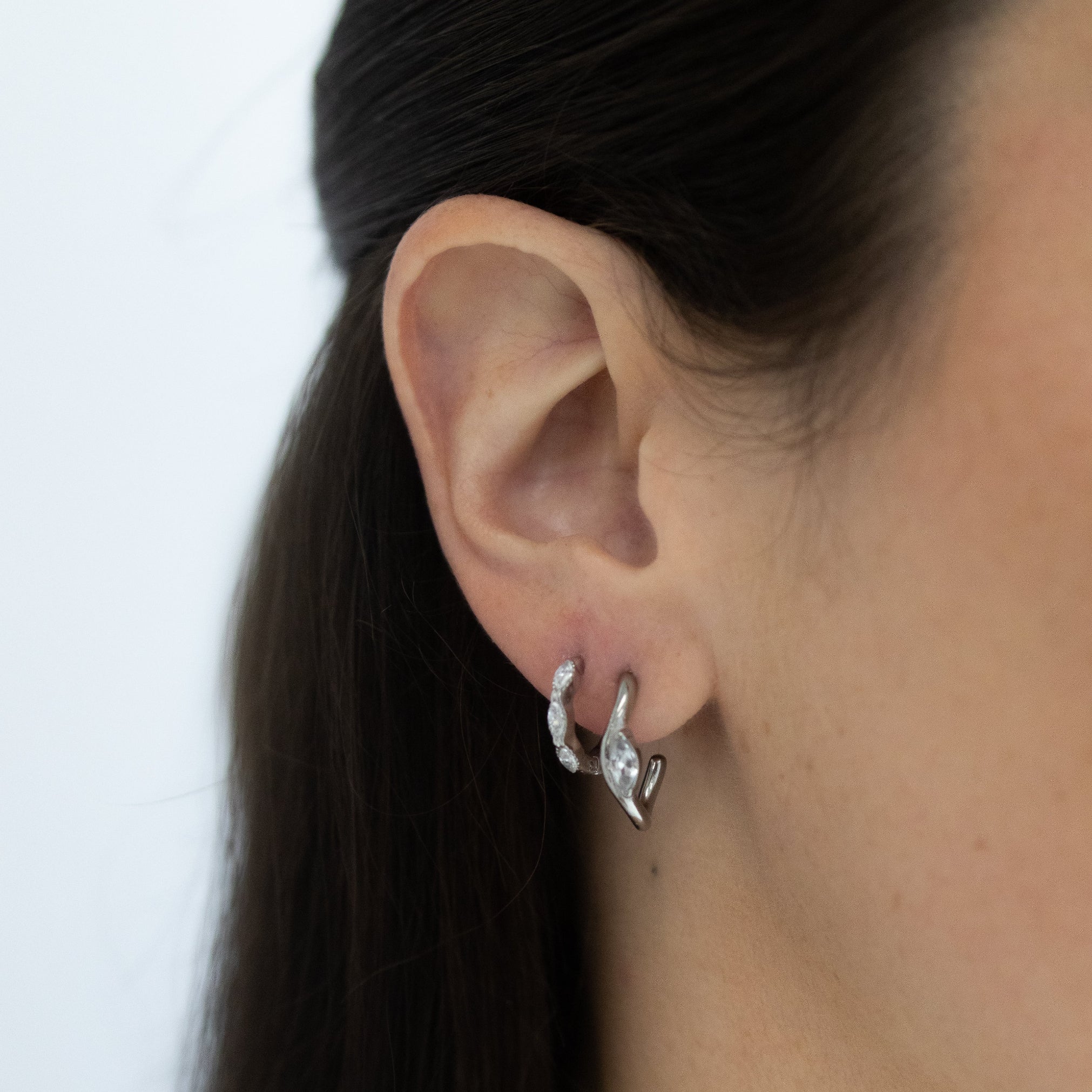 A close-up of a person's ear wearing a pair of small silver huggie hoop earrings with clear stones, against a plain light background.
