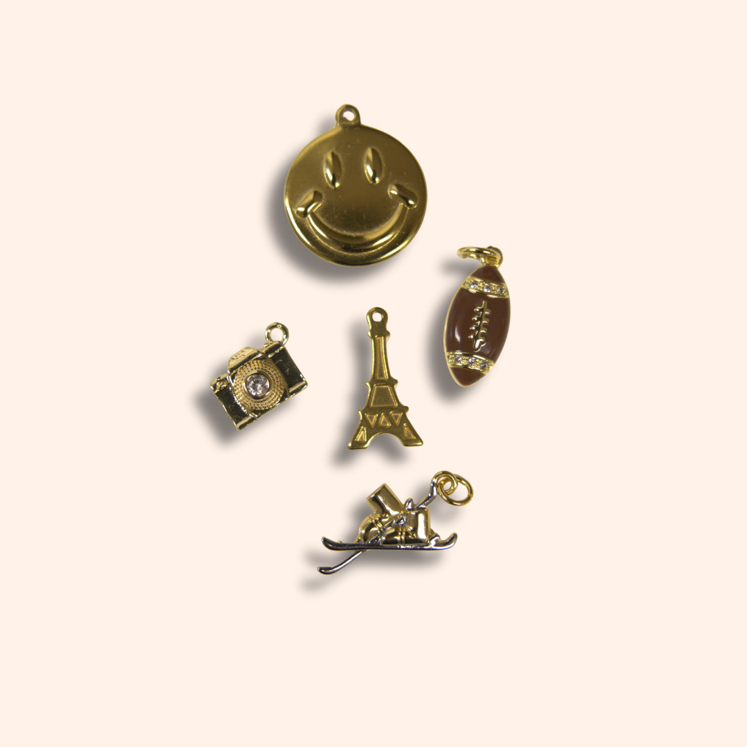 Set of gold charms including a smiley face, Eiffel Tower, camera, and shoe on a beige background