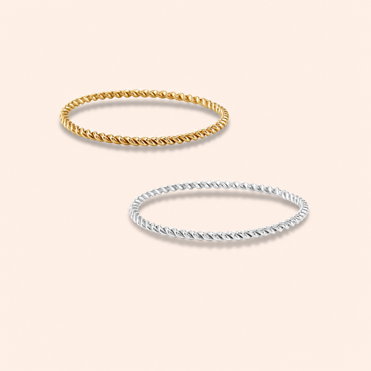 Two twisted rope-style bangles, one in yellow gold and one in silver, displayed on a light peach background.
