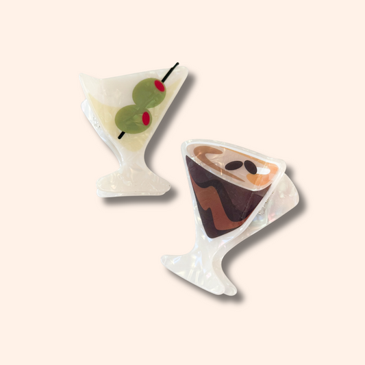 Two cocktail-themed resin pins shaped like a martini glass with olives and a chocolate milk cocktail, displayed on a solid pale peach background.