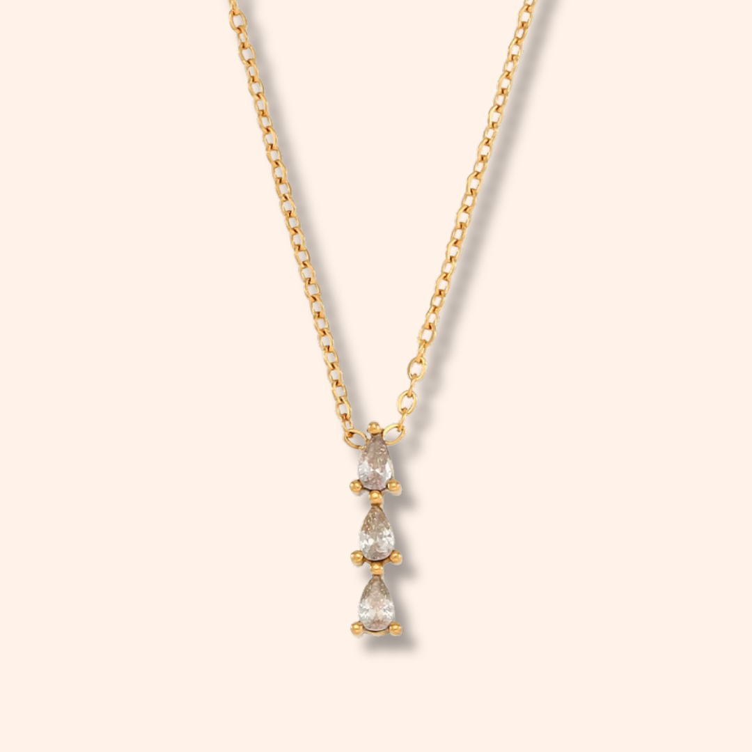 A delicate gold chain necklace featuring a vertical drop pendant with three pear-cut clear gemstones in a gold setting, displayed against a solid pale cream background.
