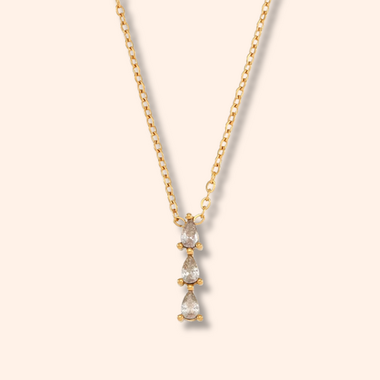 A delicate gold chain necklace featuring a vertical drop pendant with three pear-cut clear gemstones in a gold setting, displayed against a solid pale cream background.