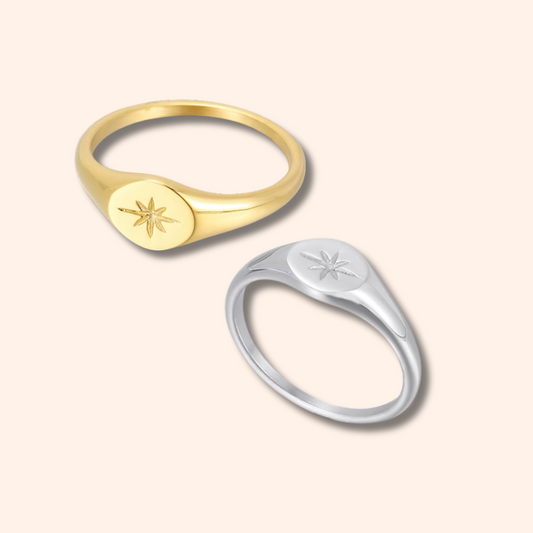 Two signet rings with oval starburst engravings, one in yellow gold and one in silver, displayed on a solid pale peach background.