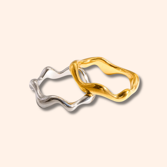 Two abstract wavy open ring earrings in polished yellow gold and silver, overlapping on a light cream background.