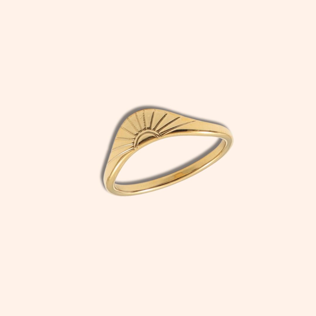A gold ring with a wavy, sunburst-inspired top design, shown on a solid pale peach background.