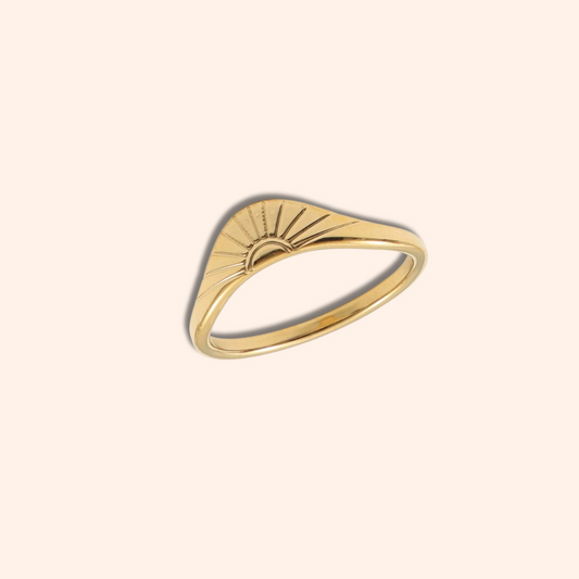 A gold ring with a wavy, sunburst-inspired top design, shown on a solid pale peach background.